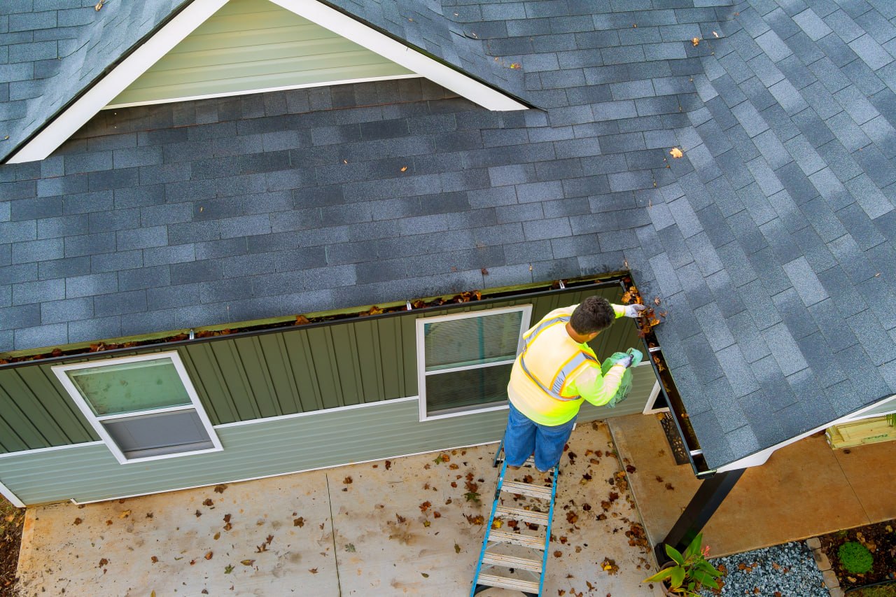 Worker cleaning leaves from gutters on a residential asphalt shingle roof